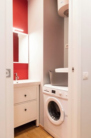 Une petite buanderie équipée d’un lave-linge, d’un évier et d’un miroir.