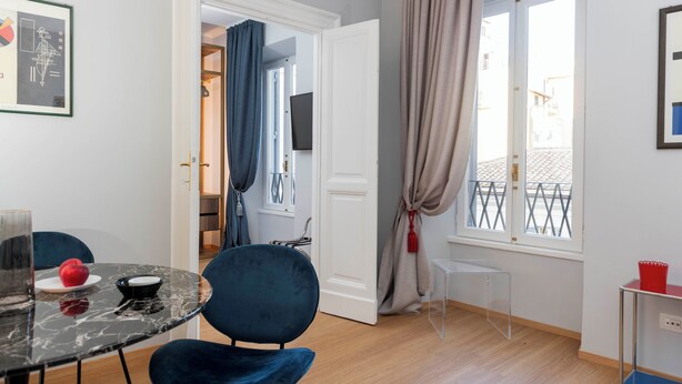 A room with a dining table, chairs, a clear glass partition, a TV, and a window with curtains. - Design Apartment in Spanish Steps (Roma)