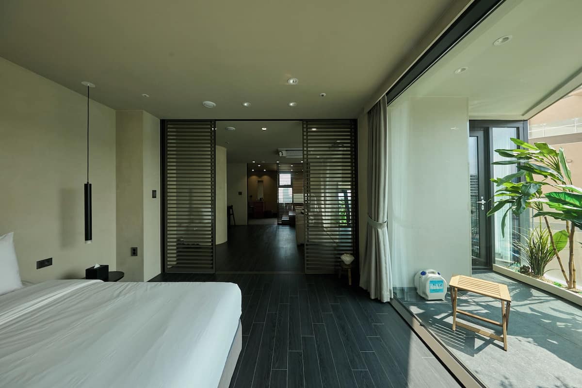 Quarto de hotel moderno com uma cama grande, uma mesinha, uma planta e vista para a cidade.