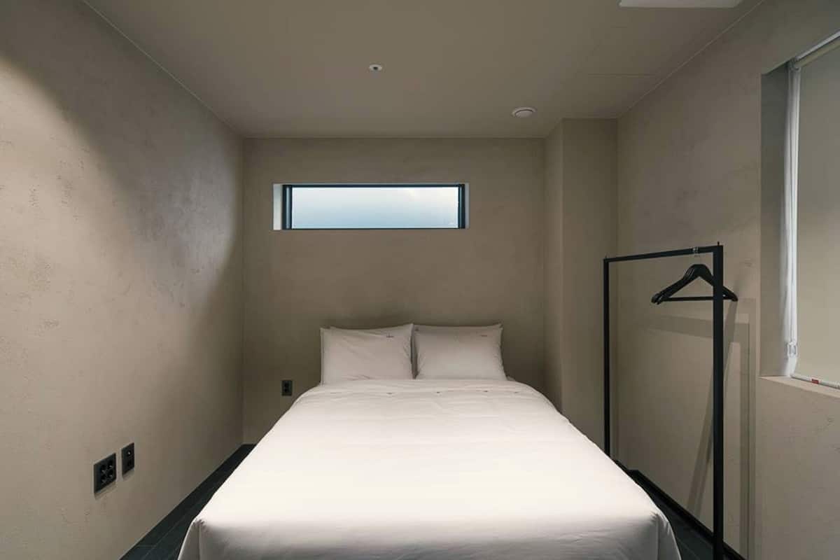 Quarto de hotel estreito com uma cama de solteiro, uma janela e um cabideiro.