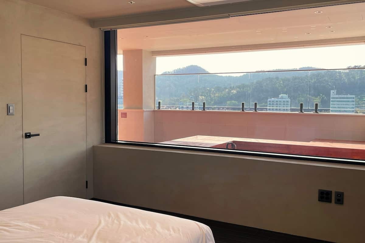 Quarto de hotel com uma grande janela, uma cama e vista para a paisagem urbana e montanhas.