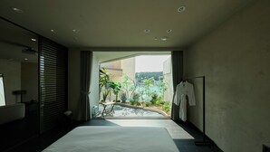 Une chambre d’hôtel moderne avec un grand lit, une salle de bain avec un miroir et une vue sur la verdure et un bâtiment, le tout visible à travers une porte ouverte.