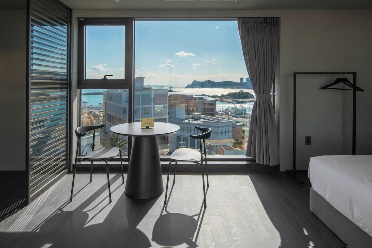 Quarto de hotel com uma janela grande, uma cama, uma mesa redonda com duas cadeiras e um cabideiro.
