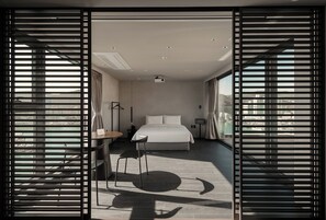 Une chambre d’hôtel moderne dotée d’un grand lit, d’un bureau avec une chaise, d’un lampadaire et offrant une vue sur la ville grâce à de grandes fenêtres du sol au plafond.