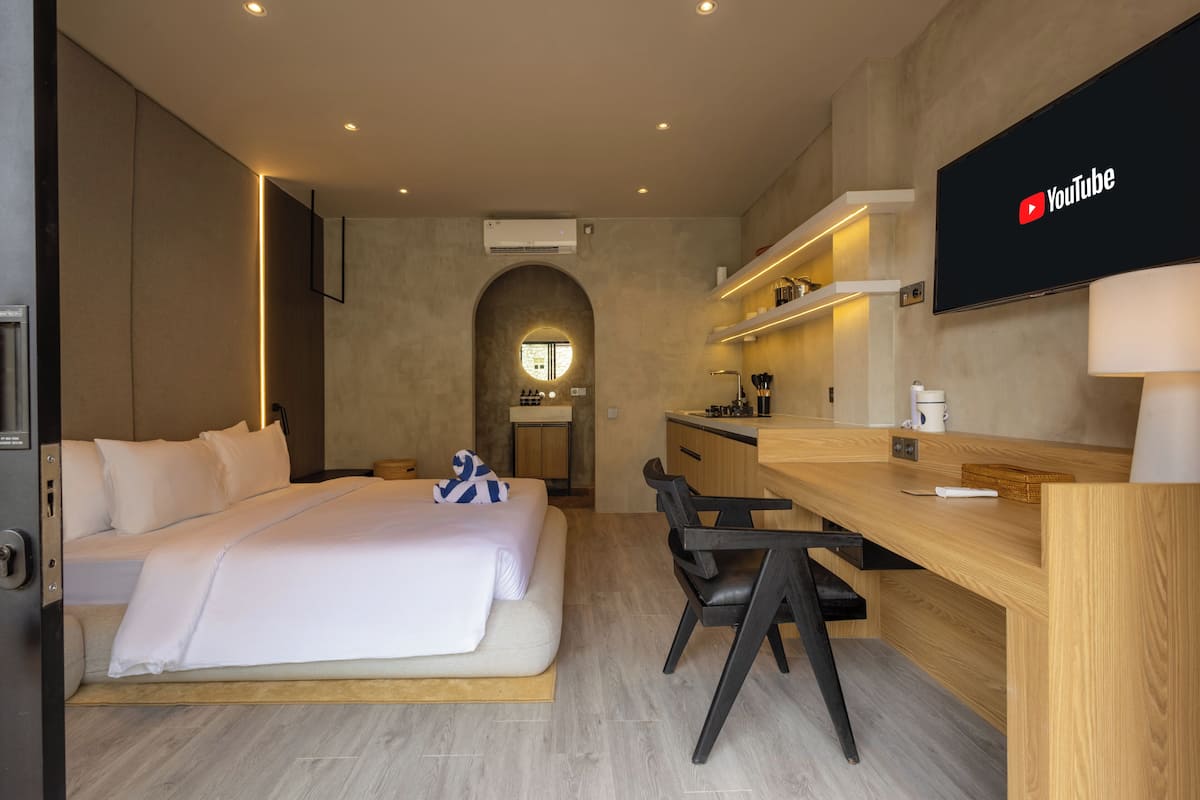 A modern hotel room with a bed, desk, chairs, a TV, and a kitchen area.