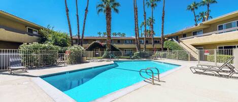 A swimming pool with palm trees, a fenced area, and a building with balconies.