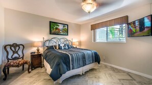 Een slaapkamer met een bed, een stoel, een nachtkastje, een lamp, een schilderij, een raam en een plafondventilator.