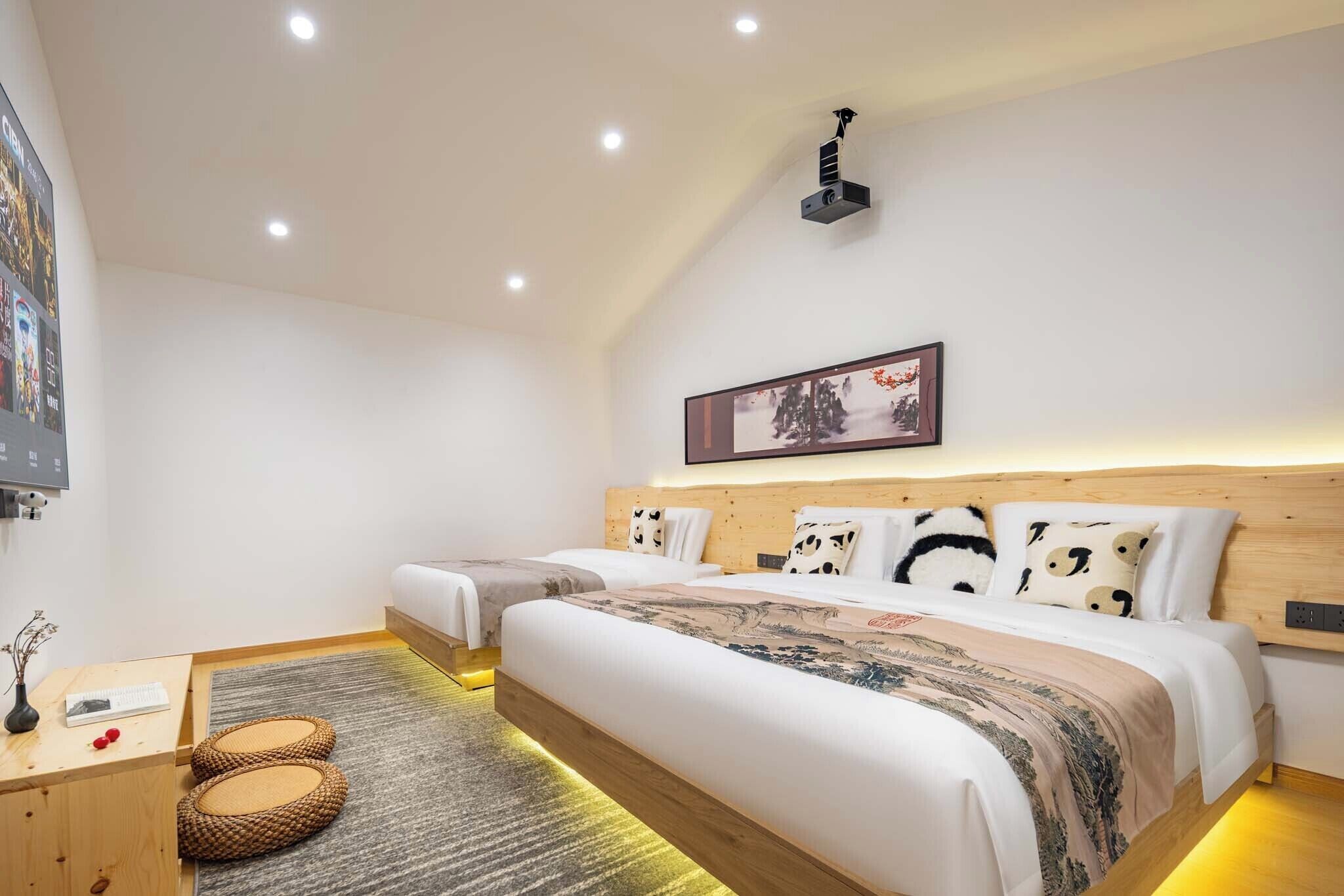 Une chambre avec deux lits, une tête de lit en bois, un projecteur au plafond, un tableau encadré et une petite table sur laquelle est posé un vase.
