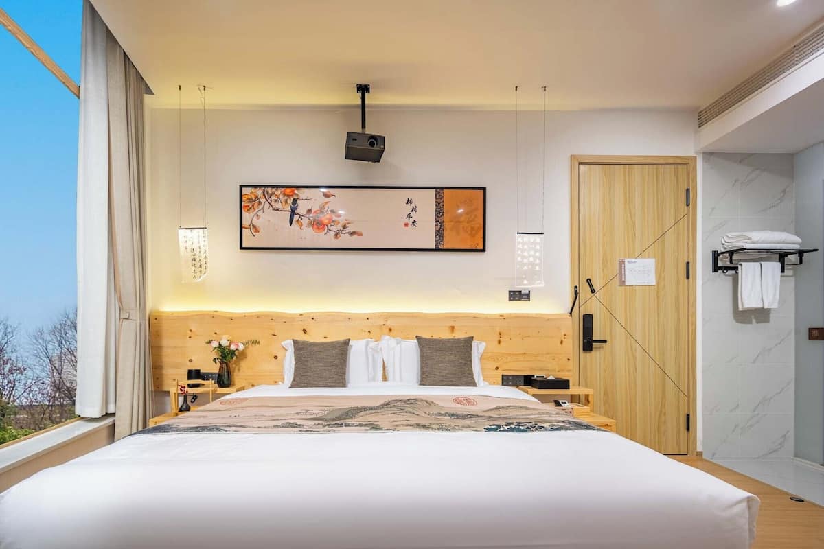 Una habitación de hotel moderna con una cama grande, cabecera de madera, un cuadro, una mesita de noche y un proyector incorporado en la pared. Se observa también una puerta con perchero.