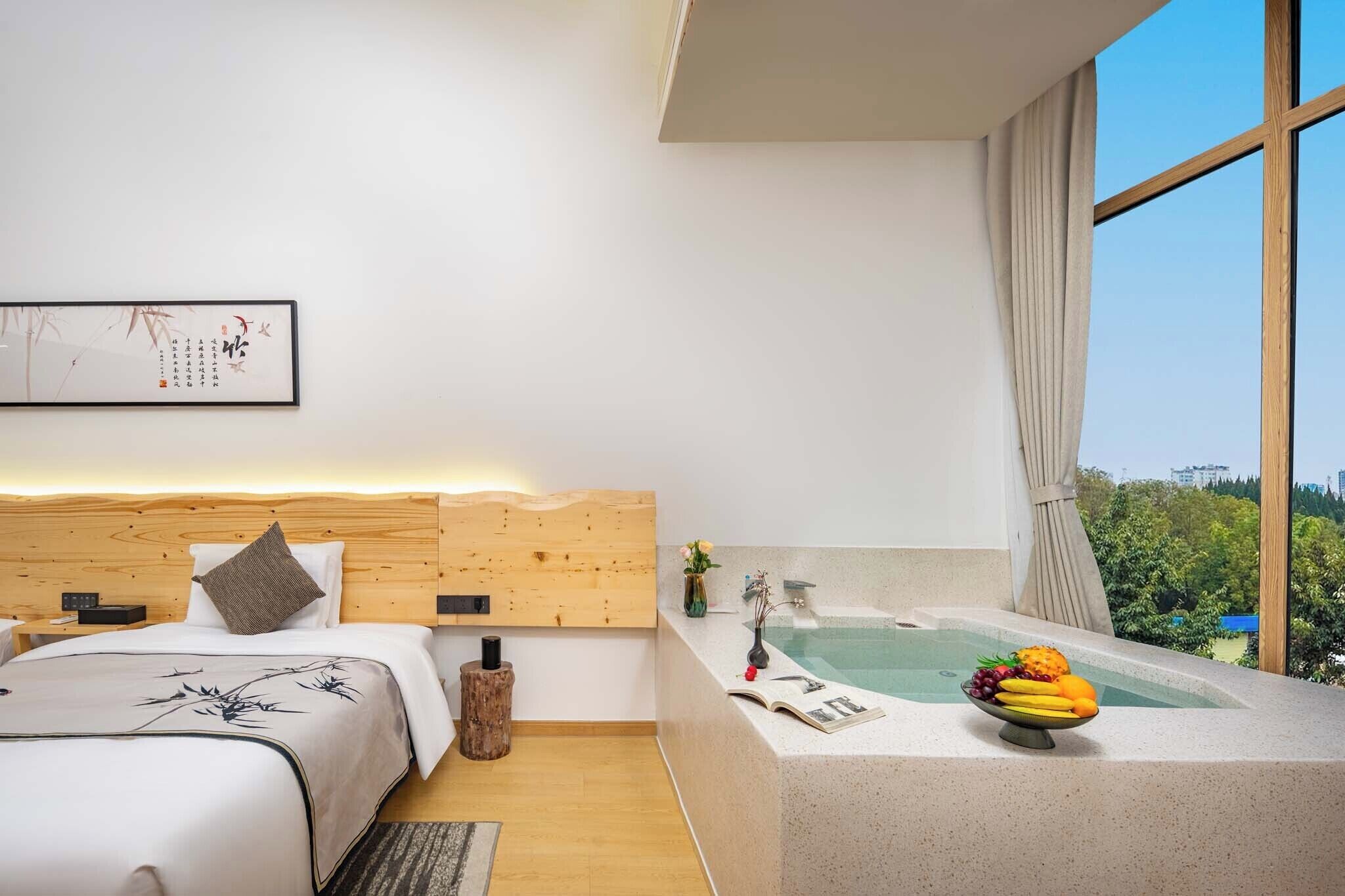 A modern hotel room with a wooden headboard, a large window, a bathtub, and a fruit bowl on the side table.