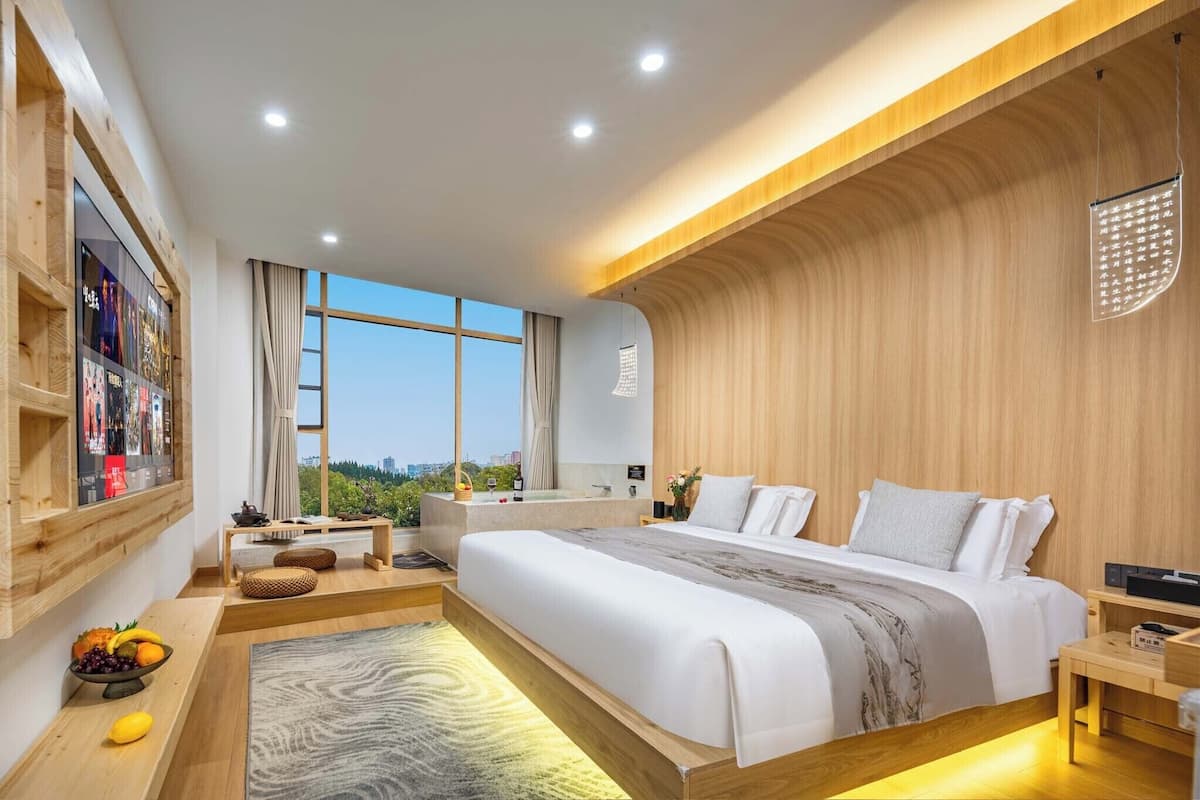 Un dormitorio moderno con una cama grande, detalles de madera, vistas a la ciudad y una pequeña mesa con frutas.