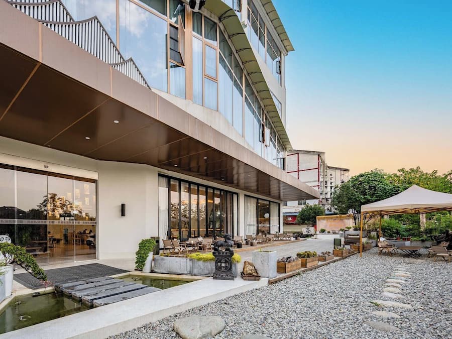 Un edificio moderno con un área de estar exterior cubierta, una pequeña fuente de agua y un sendero de grava.