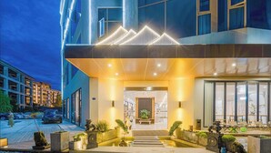 Ein moderner Hotel Eingangsbereich mit gut beleuchteter Rezeption, einer Sitzecke mit Stühlen und einem kleinen Garten mit Pflanzen und dekorativen Statuen.