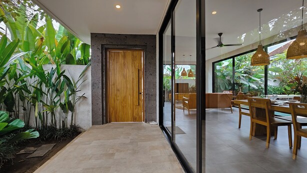 Un interno moderno con una porta in legno, un'area pranzo con mobili in legno e ampie finestre affacciate sul verde.
