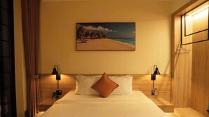 Quarto de hotel com cama, duas luminárias de cabeceira, pintura de uma praia e um guarda-roupa de madeira.