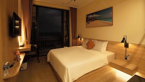 Quarto de hotel com uma cama, escrivaninha, cadeira, televisão e um quadro de uma praia.