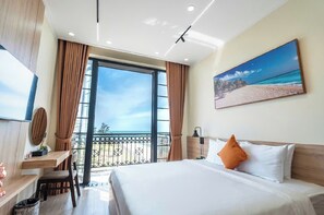 Ein Hotelzimmer mit einem großen Bett, einem Schreibtisch, einem Sessel, einem Fernseher und einem Balkon mit Blick auf den Strand.