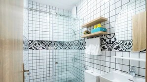 Um banheiro com azulejos brancos e pretos estampados, box de vidro, vaso sanitário branco e pia branca com prateleira de madeira acima.