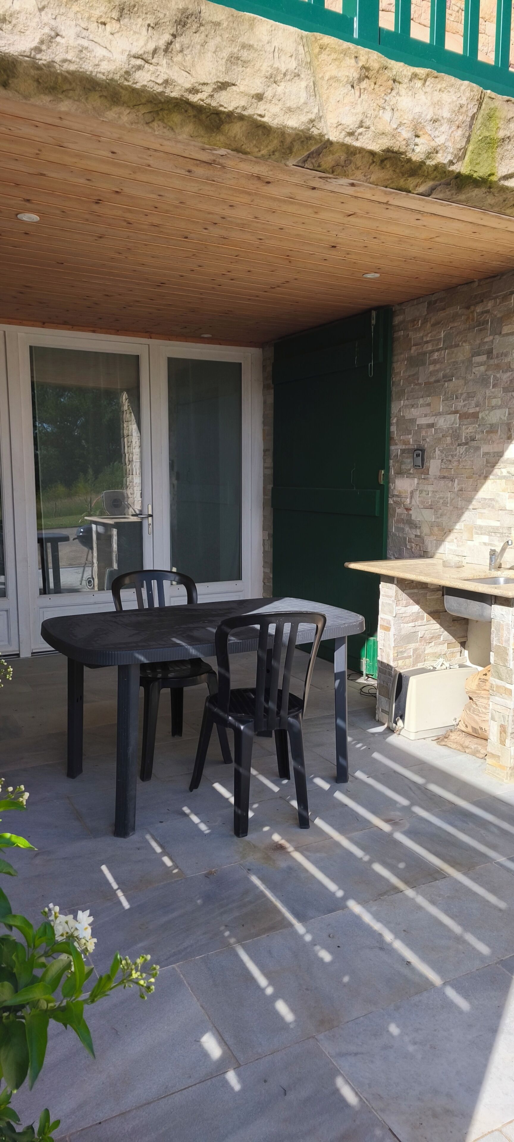 Een terras buiten met een stenen tafel en stoelen, een stenen muur en een groene deur.