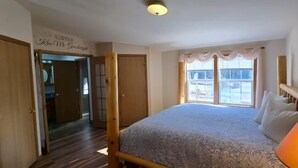 Ein Schlafzimmer mit Bett, Nachttischlampe, einem Fenster mit Vorhängen und einer Tür zu einem anderen Raum.