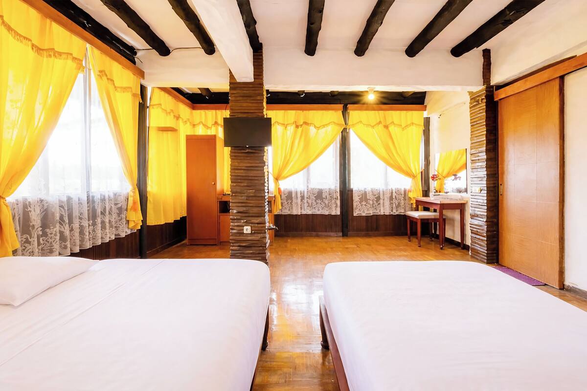 A room with two beds, a wooden desk, a stone pillar, and yellow curtains.