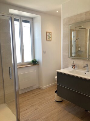 Un baño moderno con cabina de ducha de vidrio, un inodoro blanco, un tocador oscuro con espejo y una ventana con planta.