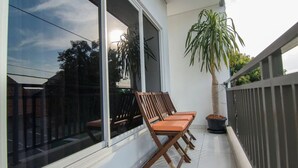 Un balcon agrémenté d’un pot de fleurs, d’une chaise en bois et d’une fenêtre donnant sur le ciel et les arbres.