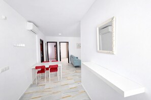 A hallway with a white wall, a white ceiling, a white floor with patterned tiles, a white table with red chairs, and a blue sofa. - Apartamento San Ignacio de Loyola, sol y ola III (Conil de la Frontera)