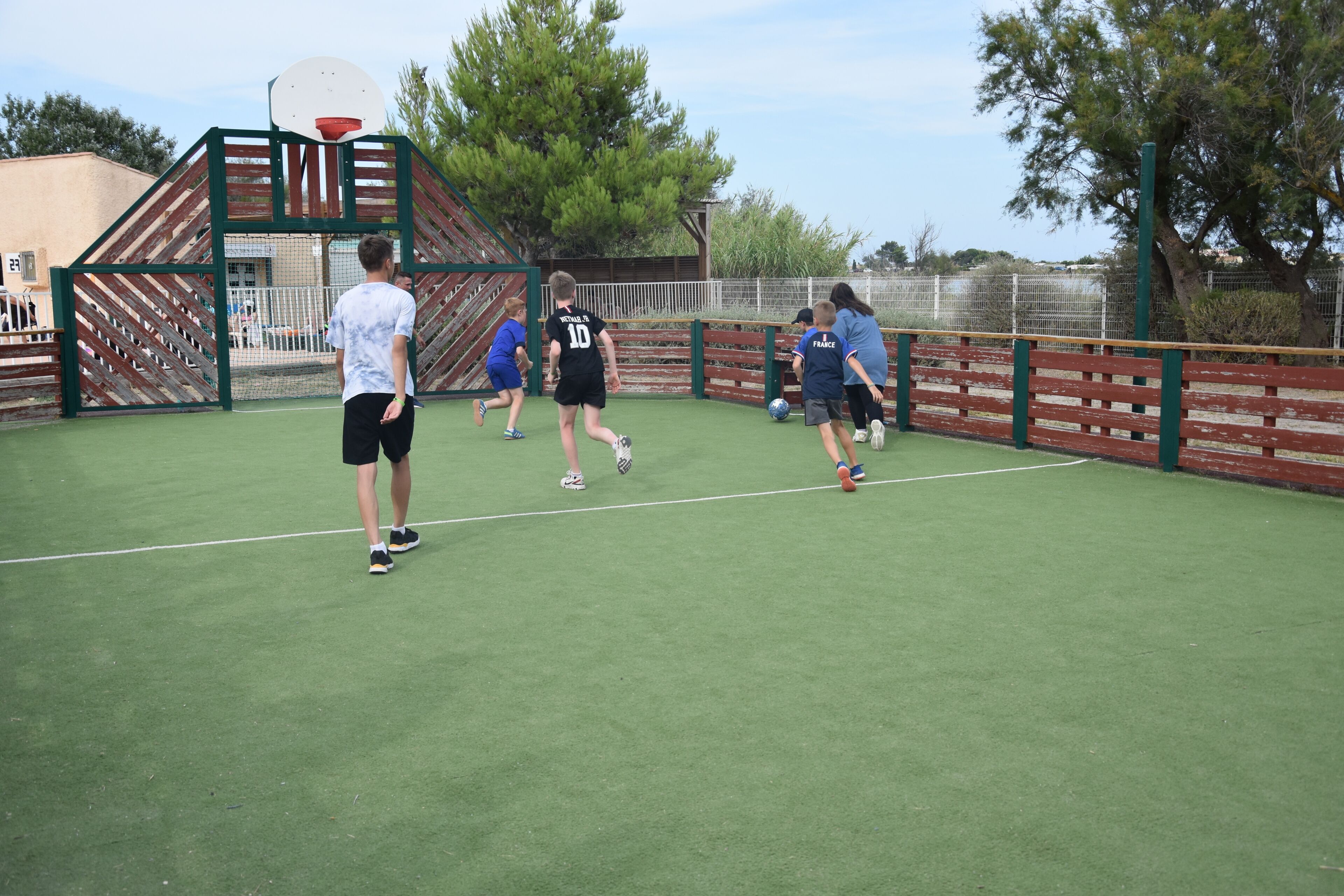 A sports court with a basketball hoop, fencing, and trees in the background.