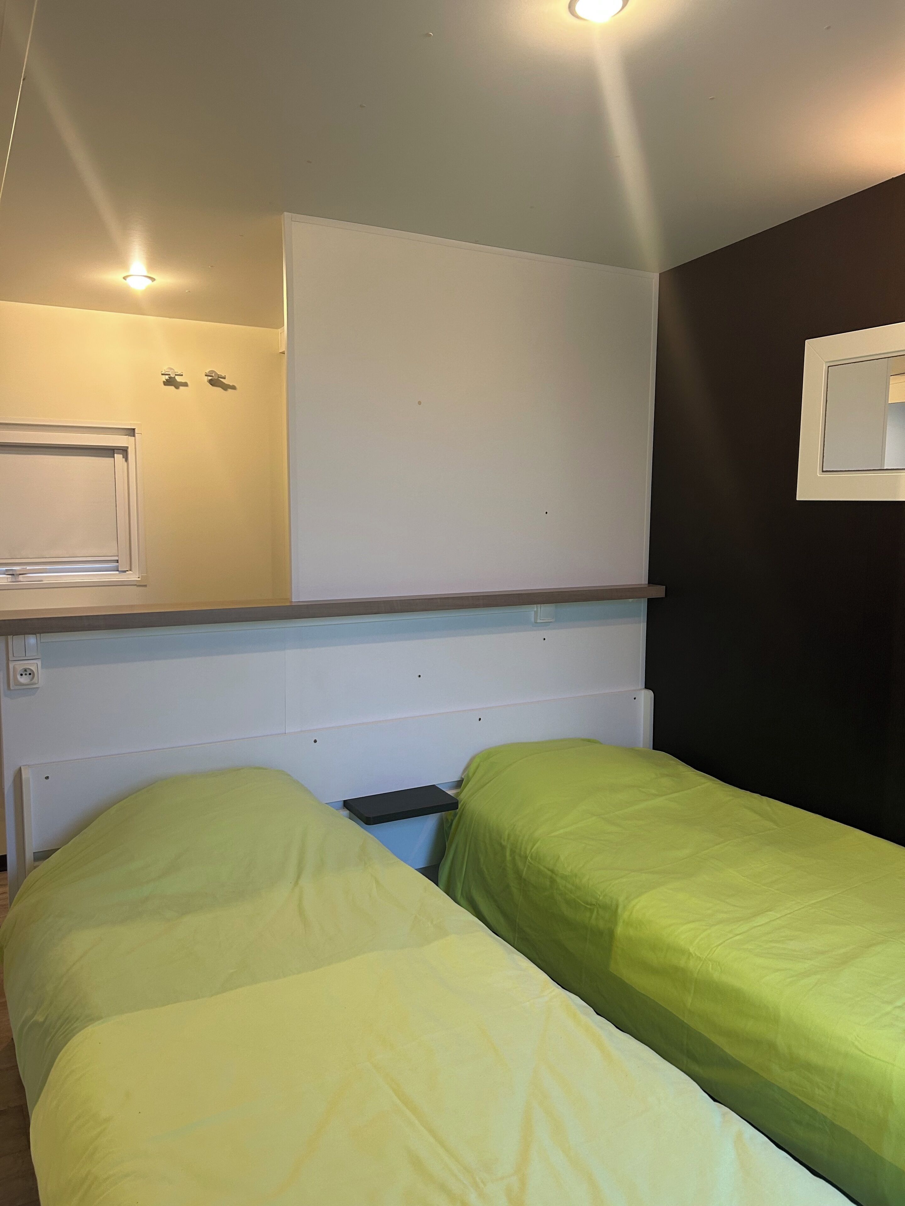 Un petit logement composé d’une seule pièce, comprenant deux lits, une fenêtre, un miroir et un luminaire au plafond.
