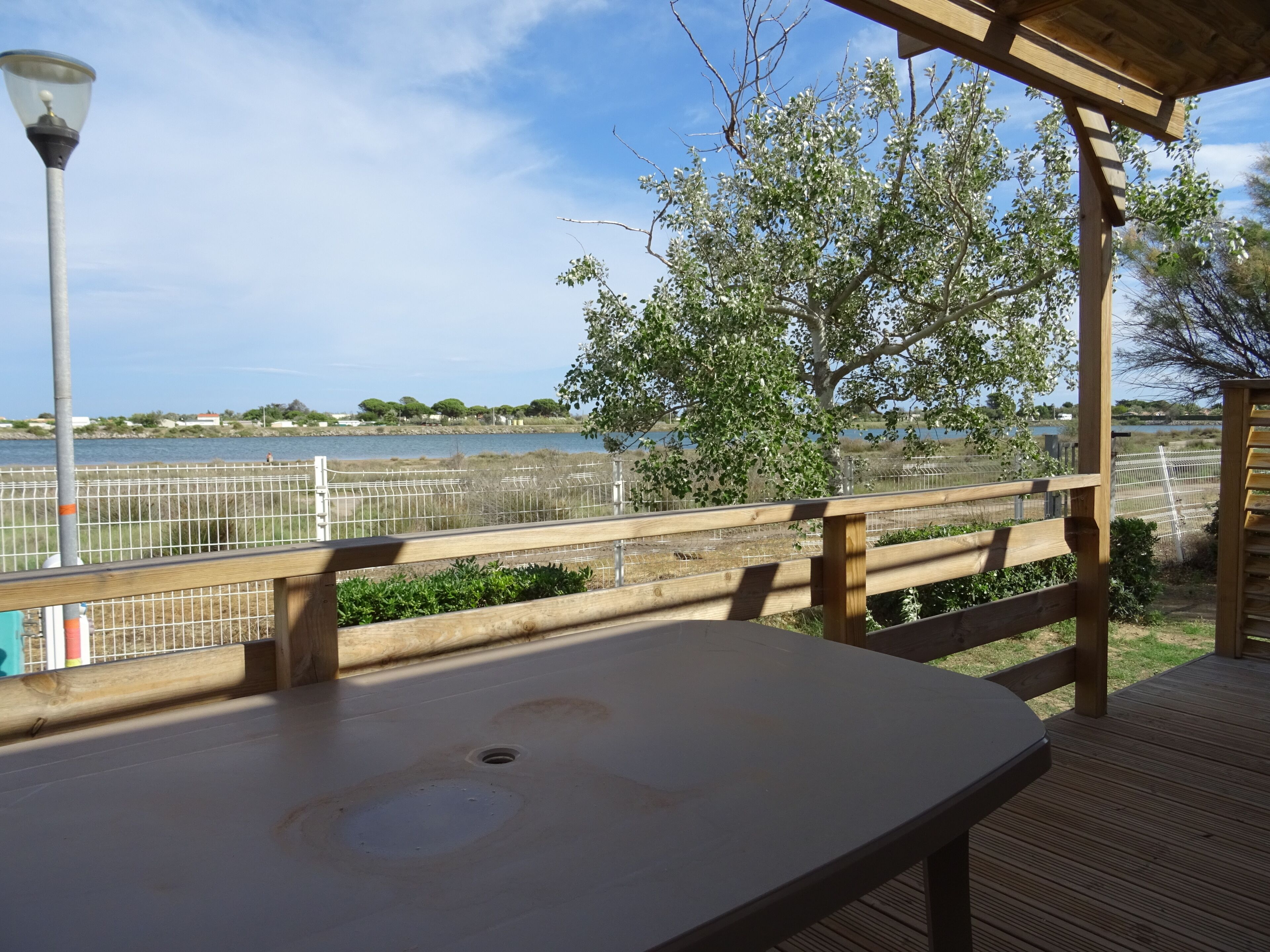 A wooden deck with a table, a lamp post, and a view of a body of water and trees.