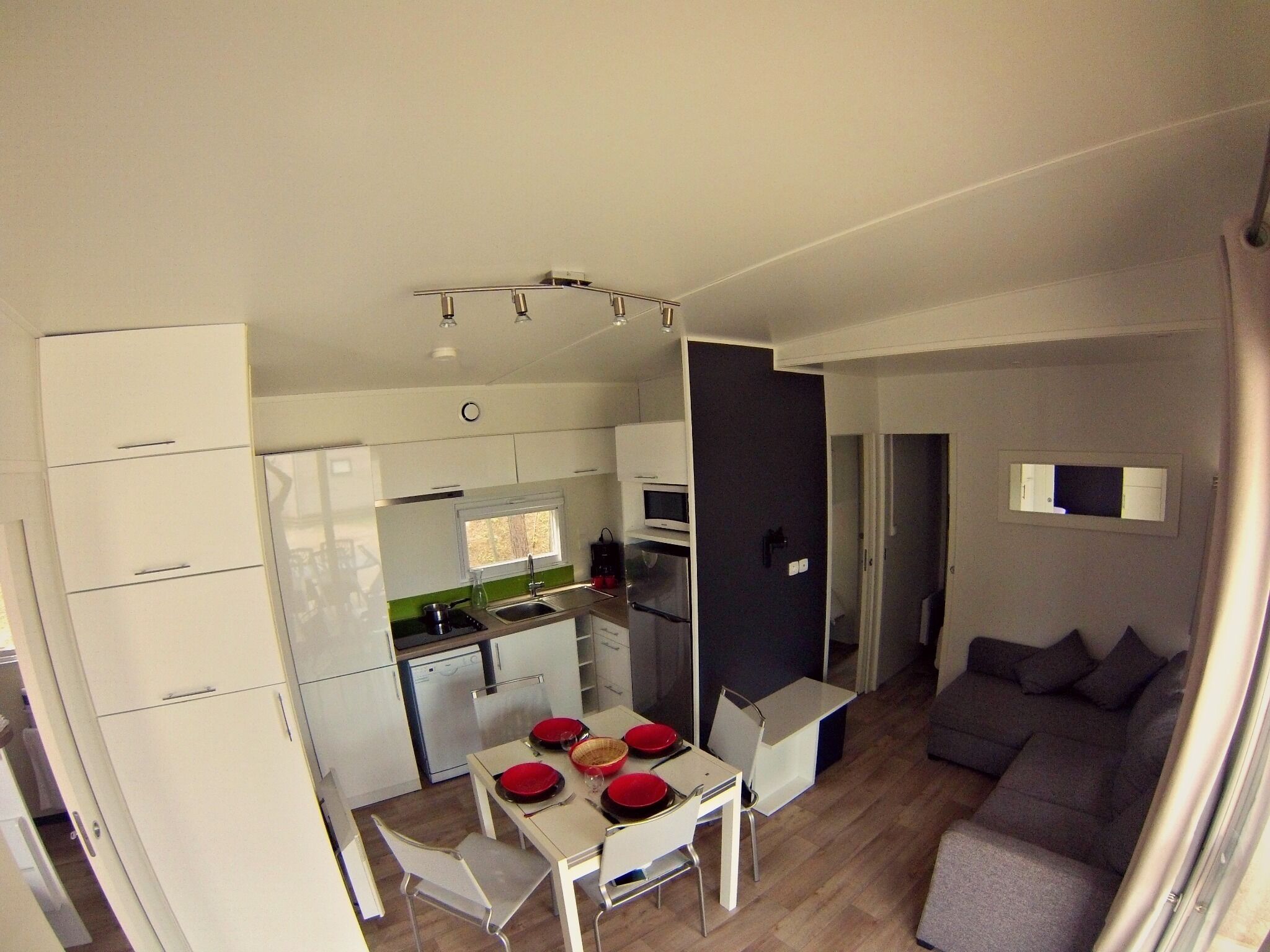 Een kleine keuken met witte kasten, een eettafel met rode borden en een grijze bank in de woonkamer.