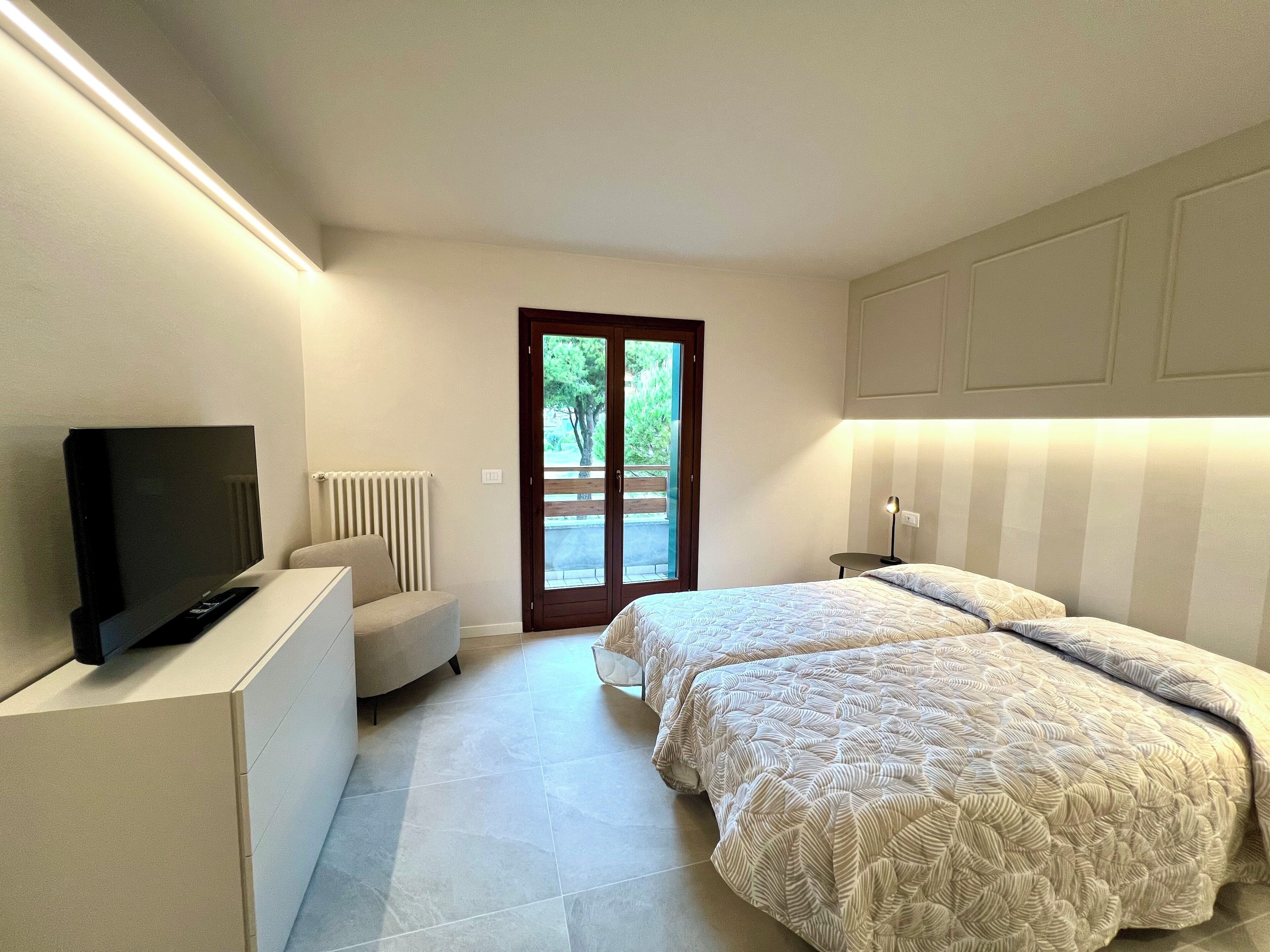 A bedroom with a TV, two beds, a chair, a radiator, and a door leading outside.