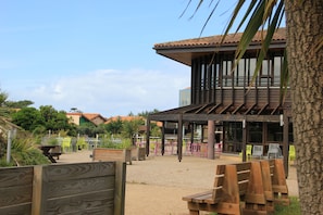 A covered outdoor seating area with wooden benches and tables, a building with a tiled roof, and a palm tree in the foreground. - Village vacances Cévéo de Mimizan-Plage  (Mimizan)