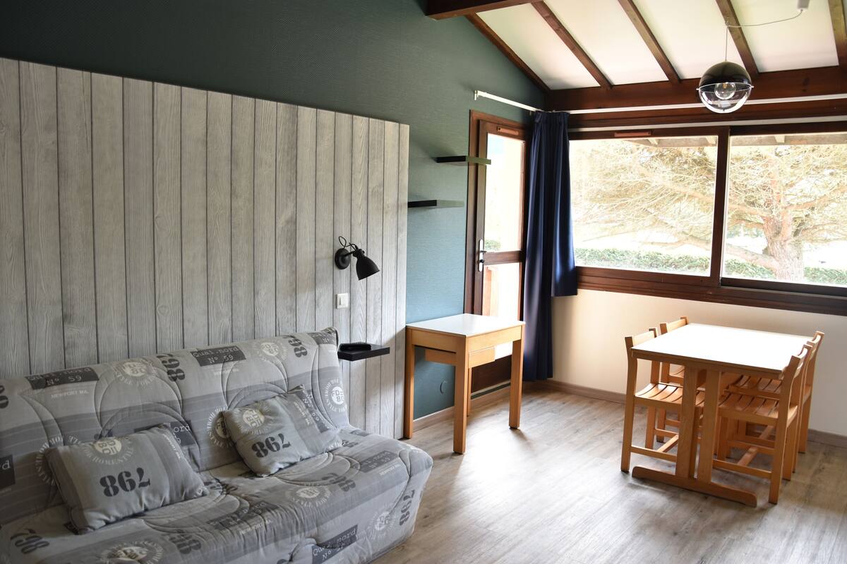 Une pièce avec un plancher en bois, un canapé agrémenté de coussins à motifs, une table en bois et une fenêtre donnant sur des arbres.