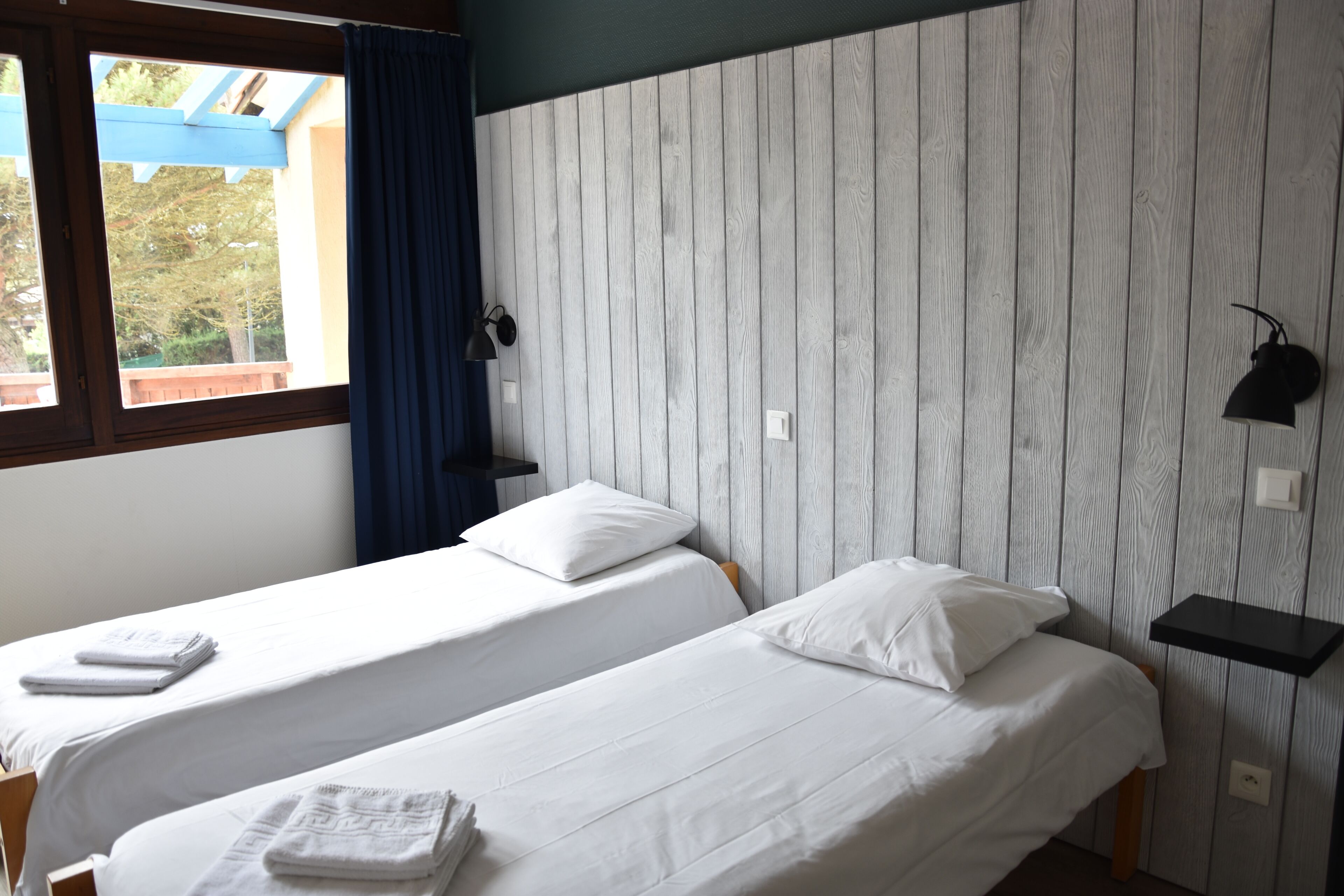 Une chambre avec deux lits simples, une fenêtre avec des rideaux, une étagère fixée au mur et une lampe fixée au mur.