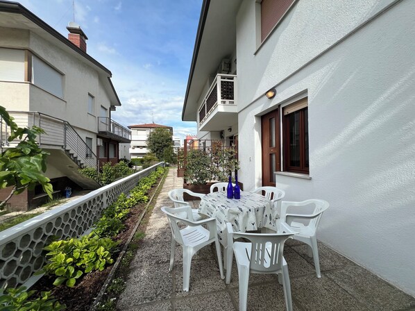 A courtyard with a table and chairs, a small garden, and a building with a balcony. - Quiet apartment with garden (Lignano Sabbiadoro)