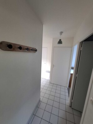 A hallway with white tiled flooring, a wooden wall-mounted shelf, a door at the end, and a hanging light fixture. - T3 furnished near the beach (Pornichet)