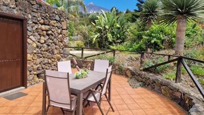 An outdoor patio with a stone wall, a wooden table with chairs, and a view of tropical vegetation and a mountain in the background. - Finca Casonas de Marengo (Icod de los Vinos)