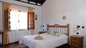 A bedroom with a wooden bed, white bedding, a wooden nightstand, a bedside table, and a window with curtains. - Finca Casonas de Marengo (Icod de los Vinos)