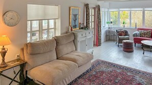 A cozy living room with a sofa, a patterned rug, a clock, and a window with blinds. - 2 bedroom cozy home in Væggerløse (Væggerløse)
