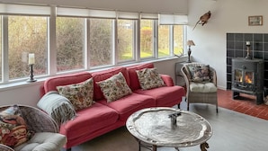 A cozy living room with a red sofa, patterned cushions, a wicker chair, a metal table, a fireplace, and large windows with a view of trees. - 2 bedroom cozy home in Væggerløse (Væggerløse)