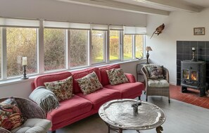 A cozy living room with a red sofa, patterned cushions, a wicker chair, a metal table, a fireplace, and large windows with a view of trees. - 2 bedroom cozy home in Væggerløse (Væggerløse)