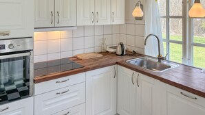 A kitchen with white cabinets, a wooden countertop, a stainless steel sink, a kettle, and a window with a view of greenery. - 2 bedroom cozy home in Væggerløse (Væggerløse)