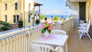 En balkon med et bord og stole, en blomstervase og udsigt til havet og bygninger.