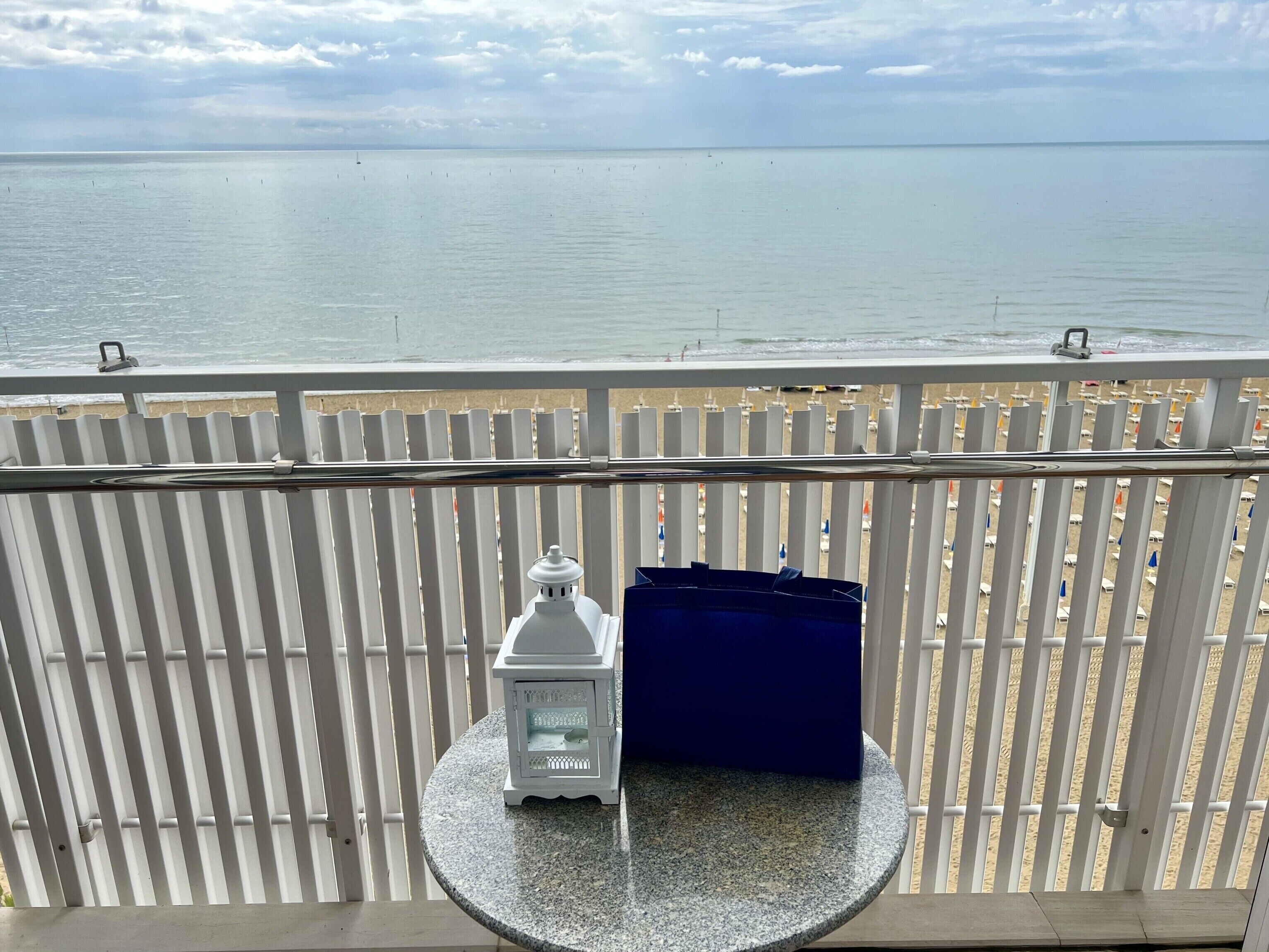 Un balcon avec une balustrade blanche, une petite table sur laquelle se trouve une lanterne blanche et un objet bleu, et une vue sur la mer et la plage.