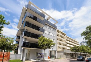 A multi-story building with balconies, a green lawn, and parked cars. - Renovated flat in Lignano center (Lignano Sabbiadoro)