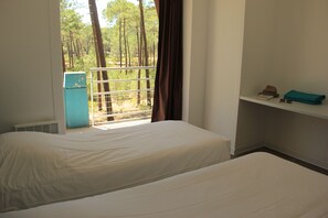 Une chambre avec deux lits, une fenêtre donnant sur des arbres, une étagère avec des objets personnels et un objet bleu sur le sol.