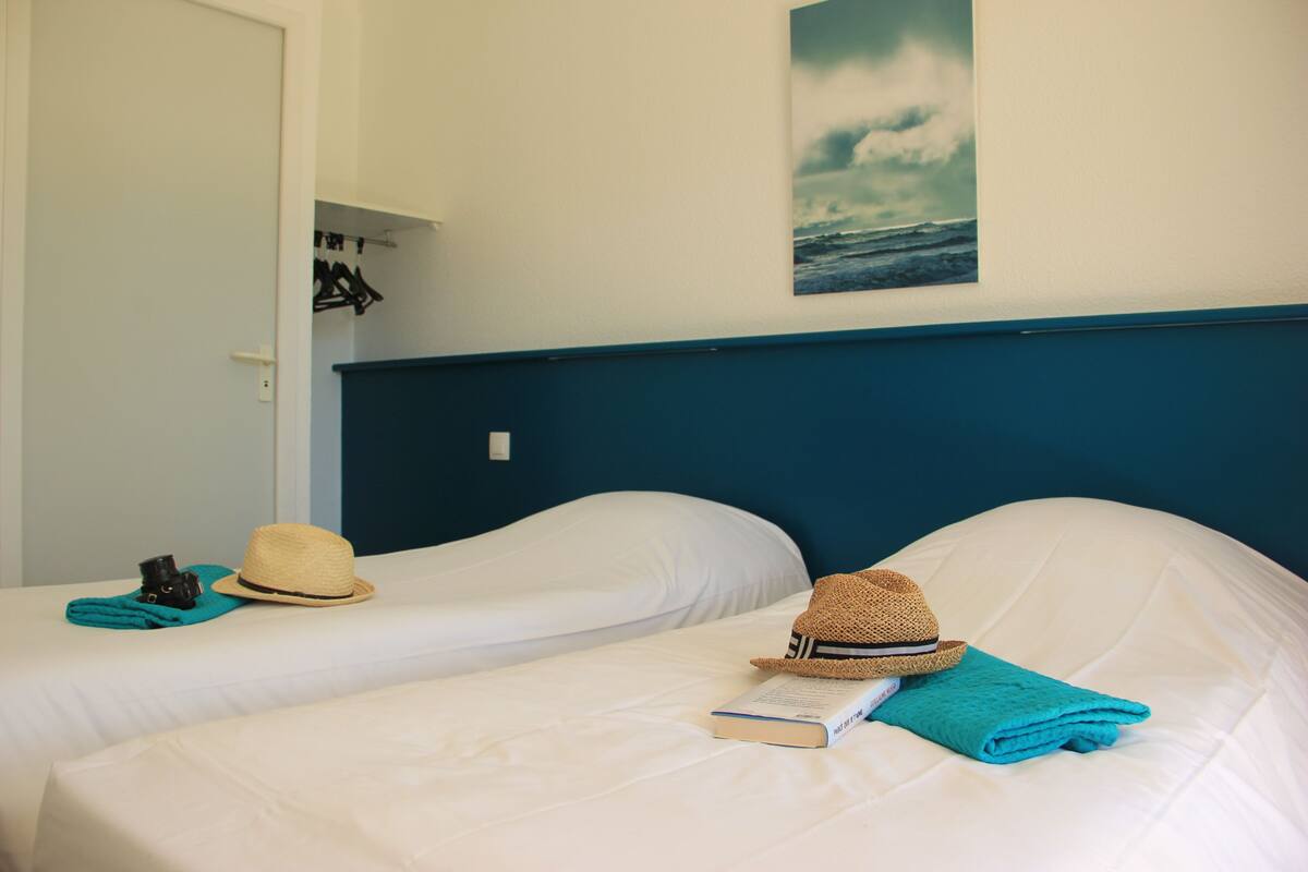 Deux lits avec des draps blancs, un chapeau, un livre et une serviette turquoise sur chaque lit.
