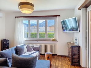 A living room with a sofa, a TV, a radiator, and a window with a view of buildings and hills.